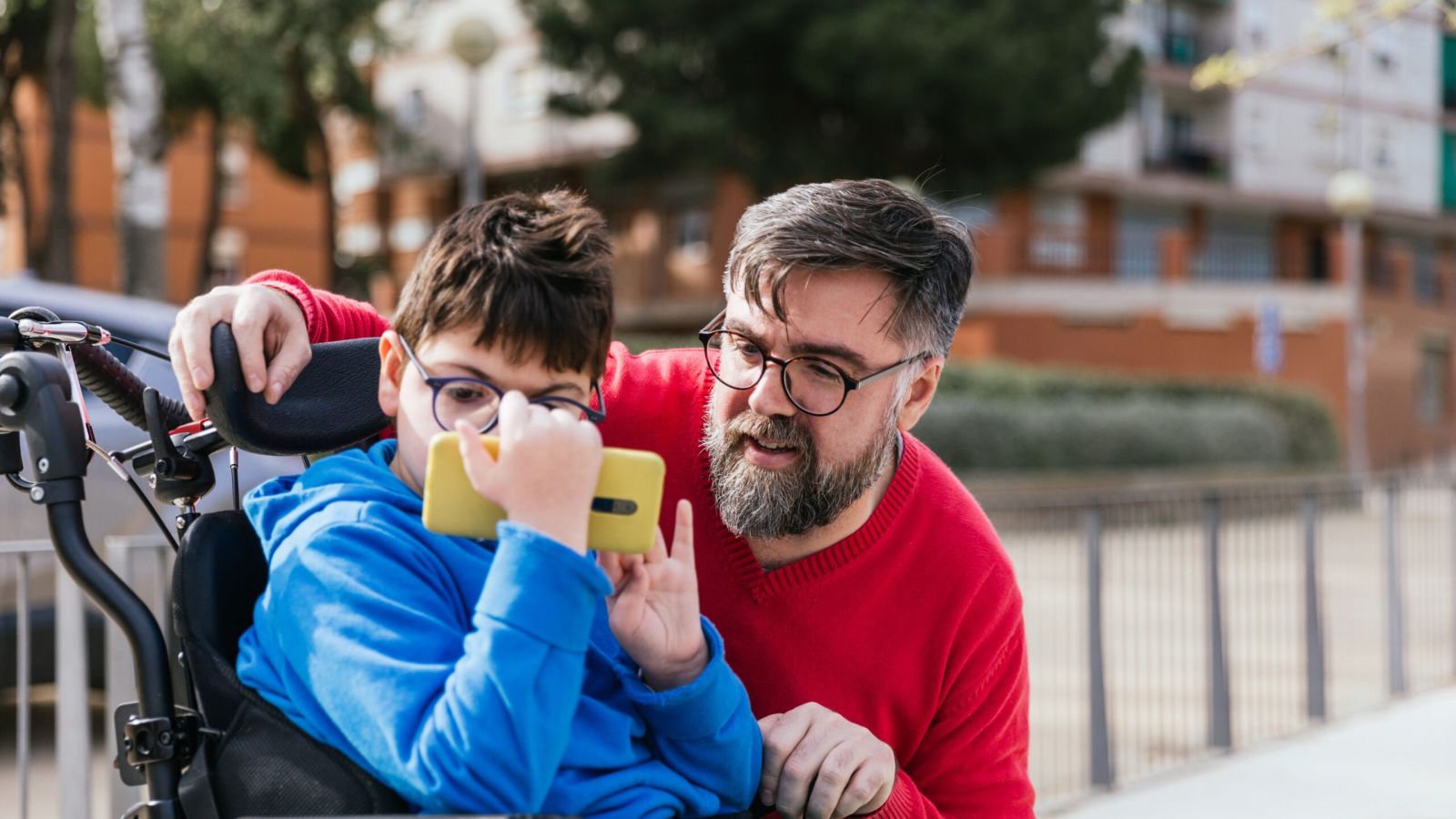 Disabled child in wheelchair watching something on the mobile phone with his father.